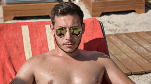 Shirtless man wearing sunglasses relaxing on lounge chair at beach during sunny day