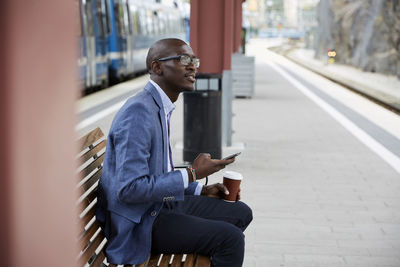 Thoughtful businessman sitting with smart phone and coffee at railroad station