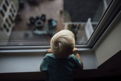 Rear view of baby boy looking through window while standing at home