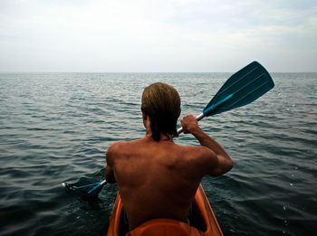 Rear view of shirtless man kayaking in sea against sky