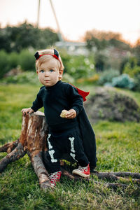 A blond boy in a devil costume with a red cloak stands near a tree stump. halloween eve