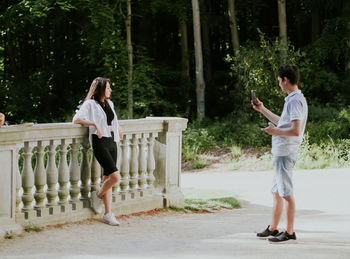 Handsome young caucasian guy photographing a girl leaning on a concrete vintage railing in a park