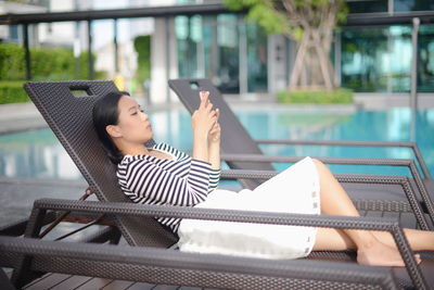 Young woman using phone while relaxing on lounge chair at poolside