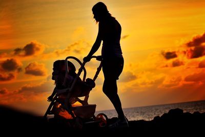 Silhouette woman pushing baby stroller at beach against sky during sunset