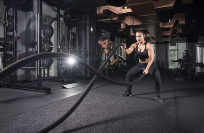 Young woman exercising with battle ropes while man in background at gym