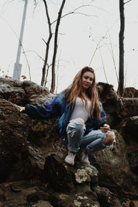 Low angle view of young woman sitting on rock formation against sky