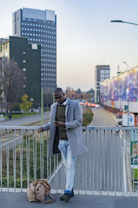 Businessman with duffel bag using smart phone leaning on railing in city