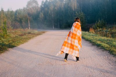 Rear view of woman wrapped in orange blanket crossing road in forest