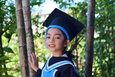 Portrait of smiling girl wearing graduation gown and mortarboard by tree trunk