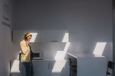 Young pregnant female in trendy striped jacket and jeans talking on smartphone while standing in modern kitchen with white furniture and microwave oven