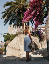 Rear view of young woman in summer dress touching beautiful pink flower in alley.