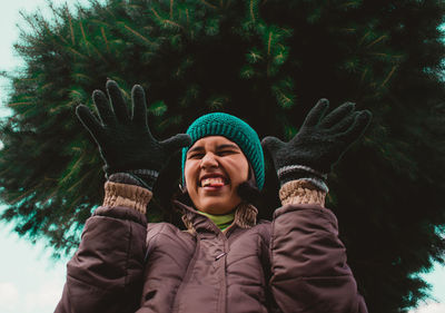Low angle view of woman making face while gesturing against tree