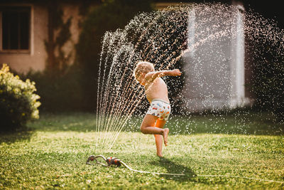 Young white boy running under the water from the sprinkler in garden