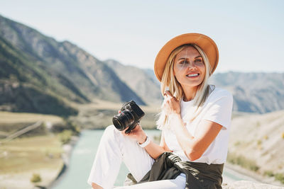 Beautiful smiling blone young woman traveler in felt hat with photo camera trip to mountains