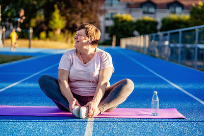 Relaxed senior plus size woman with earphones sitting on yoga mat on sports ground outdoors resting 