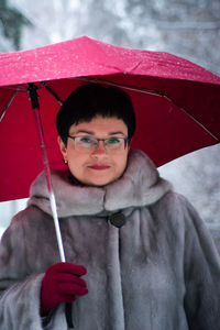 Woman in a gray fur coat with a red umbrella stands on a white, snow-covered background.