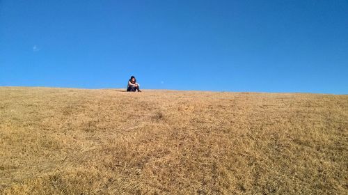 Woman sitting on grassy field against clear blue sky during sunny day