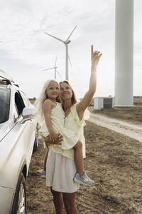 Smiling woman gesturing and standing with daughter by car at wind park