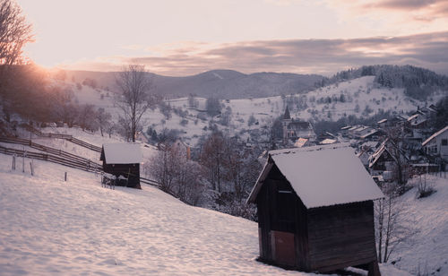 Wooden barns and cabind on snow covered field by buildings against mountains and sky