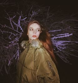 Beautiful young woman standing against tree at night during winter