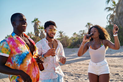 Positive young ethnic african american men and woman in casual clothes and sunglasses with beer bottle in hand proposing toast and smiling while spending time on sandy seashore with cheerful multiracial friends