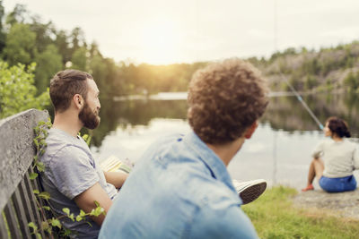 Men sitting on wooden bench while female friend fishing at lakeshore