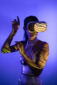 Unrecognizable young woman with long dark hair in futuristic outfit and modern vr headset exploring virtual reality standing against blue background with projector lights