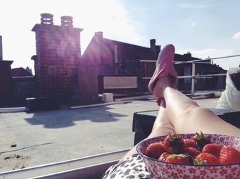 The legs and feet of a woman with a bowl of strawberries in her lap on a rooftop