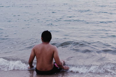 Rear view of shirtless man sitting at sea