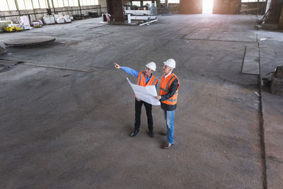 Two men with plan wearing safety vests talking in old industrial hall