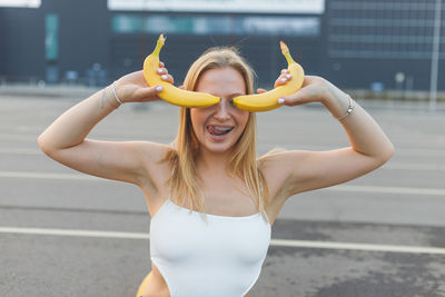 Frivolously and mischievous young woman with braces on teeth holding bananas on her face has fun