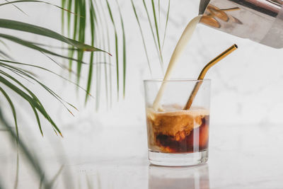 Close-up of milk poured into iced coffee glass on white marble table with metal reusable straw