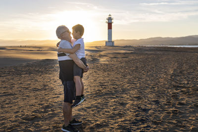 Full length of grandfather with grandson on beach against sky during sunset