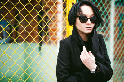 Young woman in sunglasses standing against chainlink fence