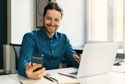 Smiling young man in casual wear looking at smartphone screen at workplace