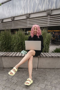 Woman with pink hair using laptop sitting on bench