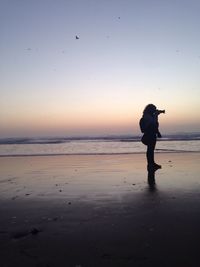 Full length of female photographer photographing at beach against clear sky during sunset