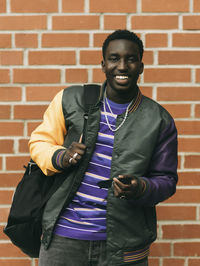 Portrait of happy young man holding sunglasses while standing against brick wall