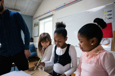 Female students writing on paper while teacher standing in classroom