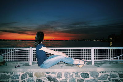 Woman sitting by sea against sky during sunset