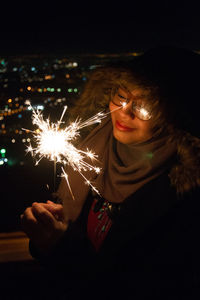 Smiling woman holding illuminated sparkler by window at night