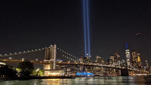 Illuminated brooklyn bridge over hudson river against sky at night with 911 tribute a light.