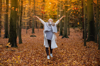 Smiling young woman with arms outstretched standing amidst falling leaves during autumn