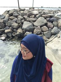 Smiling woman wearing hijab standing at sea shore against rocks