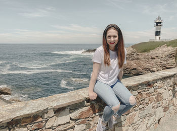Portrait of young woman sitting on rock by sea against sky