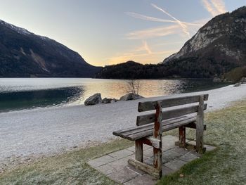 Bench by lake against sky during sunset