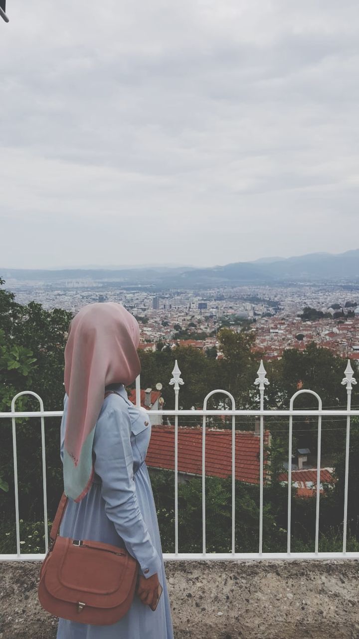 one person, real people, sky, leisure activity, lifestyles, nature, day, railing, architecture, rear view, clothing, cloud - sky, plant, casual clothing, women, tree, standing, three quarter length, environment, outdoors, looking at view