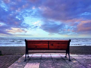 Empty bench on beach against sky during sunset