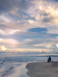 Man fishing while sitting at beach against sky during sunset