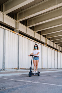 Full body of female teenager in sunglasses and protective mask riding electric scooter on paved parking lot near urban building